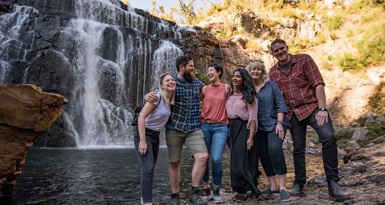 Des voyageurs souriants posent ensemble devant une cascade au milieu de rochers escarpés et de la brousse.