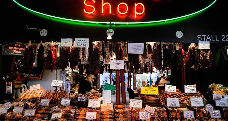 Étal de marché intérieur avec des enseignes au néon présentant des rangées de charcuteries, saucisses et collations tandis qu'un vendeur sert les clients.