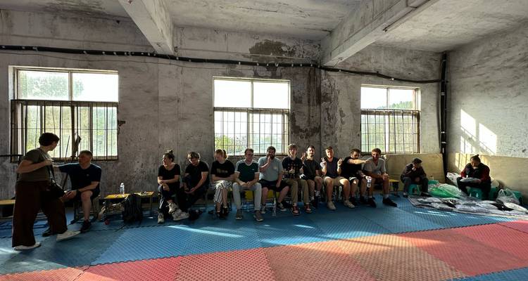 Gran grupo de estudiantes descansan en bancos dentro de un salón de entrenamiento de artes marciales luminoso con colchonetas acolchadas en el suelo.