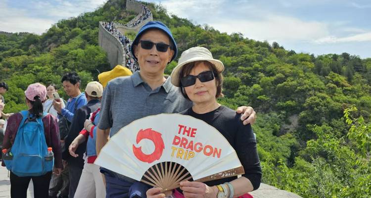 Pareja sonriente sostiene un abanico de recuerdo mientras están de pie en la Gran Muralla con colinas frondosas detrás.