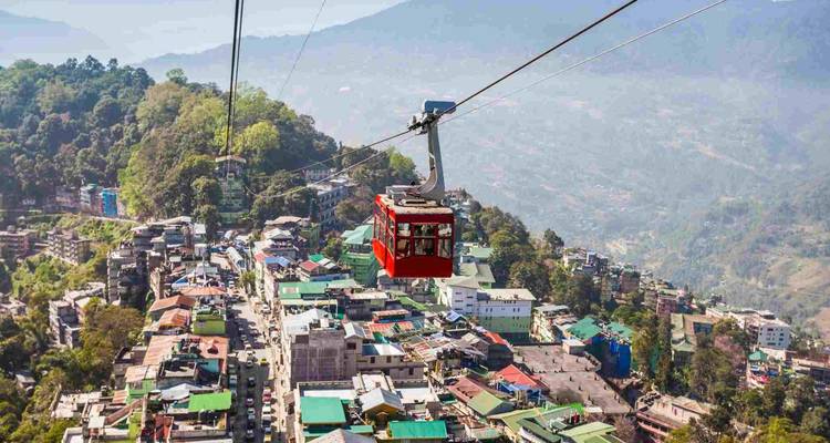 Rote Seilbahn gleitet hoch über bunter Hügelstadt mit Panorama-Bergblick.