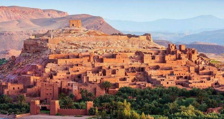 Ancien village fortifié de casbahs en terre perché sur une colline avec des montagnes arides derrière.