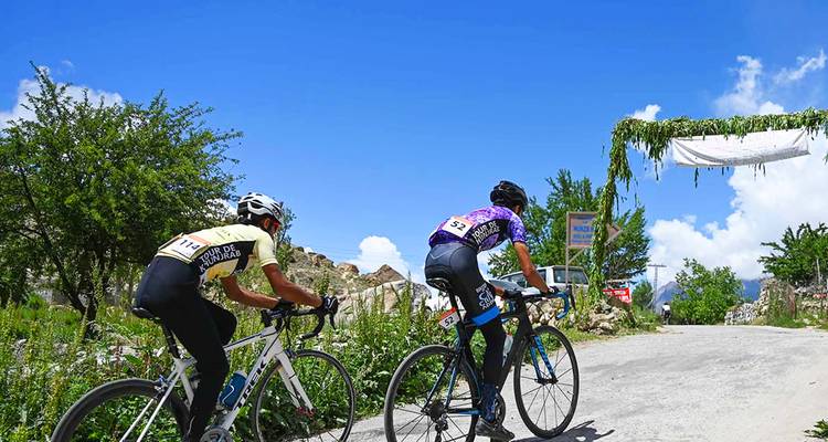 Two competitive cyclists climb a sunlit mountain road beneath a vivid blue sky.