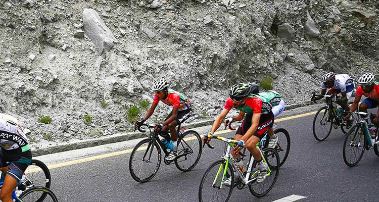 Cyclists in colorful jerseys speed along a winding highway cut through rocky slopes.