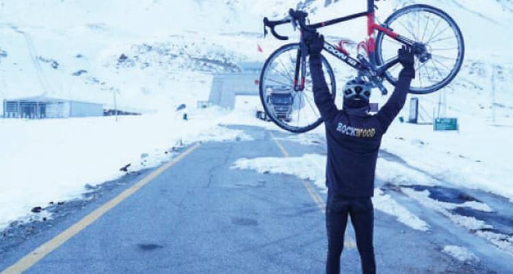 A cyclist triumphantly hoists his bike at a snow-covered high-altitude pass.