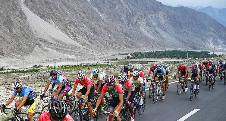 A large peloton rides through a dramatic barren valley bordered by towering peaks.