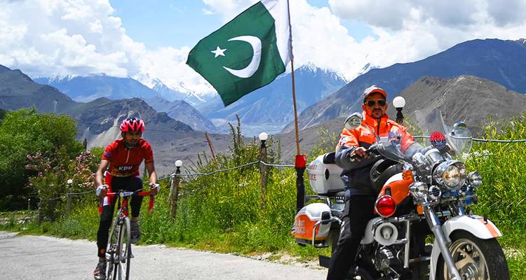 A cyclist passes a motorbike escort beneath the Pakistani flag with snow peaks in the distance.