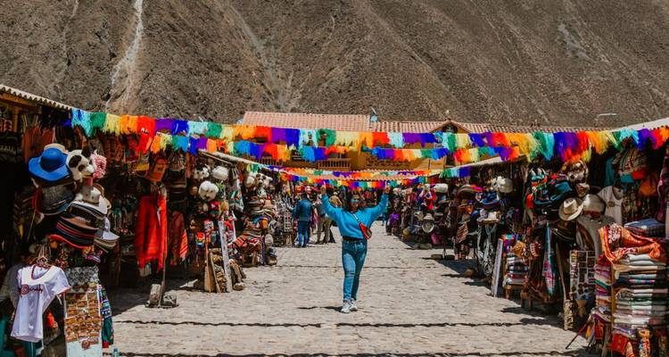 Rue de marché artisanal colorée bordée de textiles et de chapeaux avec un visiteur joyeux posant les bras écartés.