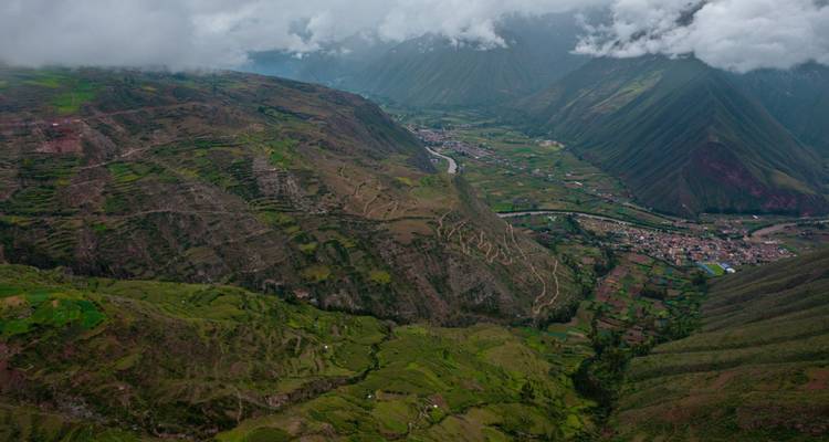 Vue aérienne sur la Vallée Sacrée avec des champs verts luxuriants, une rivière sinueuse et des sommets enveloppés de nuages.