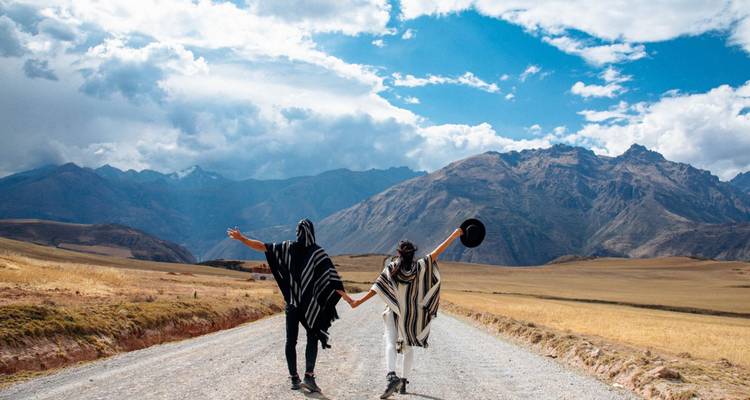 Deux voyageurs portant des ponchos se tiennent la main en marchant sur une route rurale vers les sommets andins sous des nuages dramatiques.