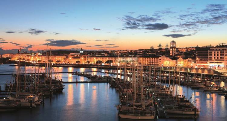 Schemerpanorama van de jachthaven van Ponta Delgada met zeilboten afgemeerd tegen een gloeiende zonsonderganglucht en verlichte waterfrontgebouwen.