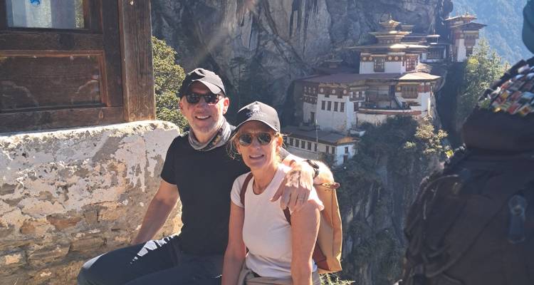 Couple assis sur un point de vue à flanc de falaise avec le Paro Taktsang visible au loin.