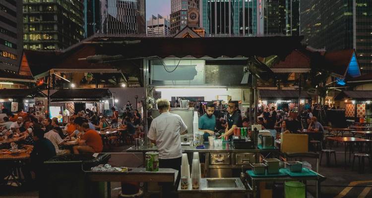 Scène nocturne animée dans un centre de restauration de rue singapourien avec des chefs préparant la nourriture et des convives remplissant les tables.
