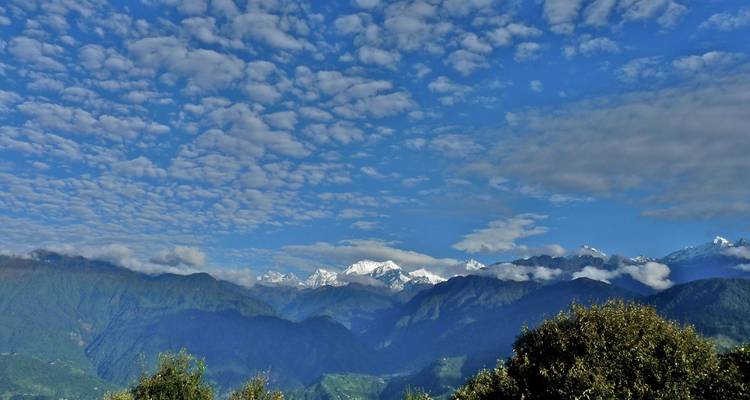 Schneebedeckte Himalaya-Gipfel erheben sich hinter bewaldeten Bergrücken unter einem weiten Himmel, der mit watteähnlichen Wolken gesprenkelt ist.