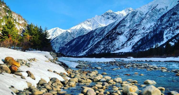 Schneegesäumter Fluss, der durch ein felsiges Tal fließt mit hoch aufragenden weißen Himalaya-Gipfeln im Hintergrund.