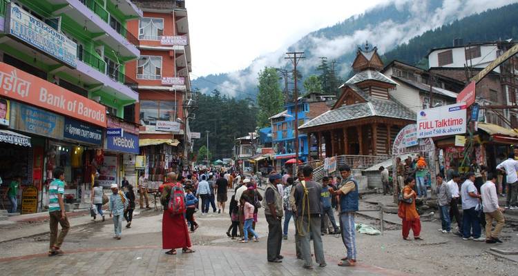 Belebte Straße in Manali gesäumt von Geschäften, Tempeln und nebligen Kiefernhügeln im Hintergrund.
