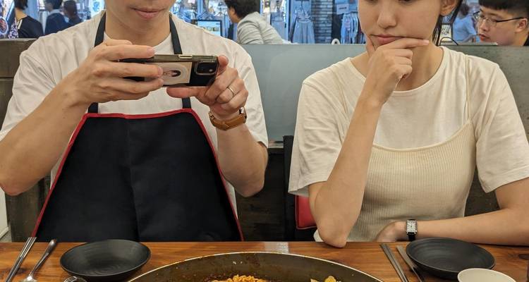 Dos comensales se sientan en una mesa de restaurante; uno fotografía la comida mientras el otro contempla el plato.