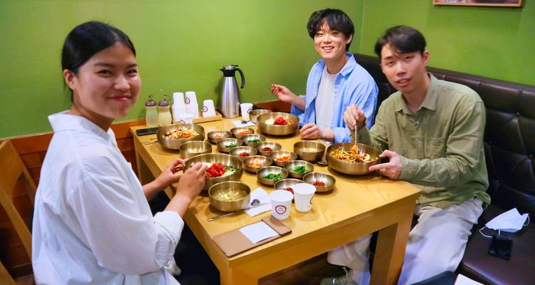 Tres amigos sonríen alrededor de una mesa cubierta con coloridos cuencos de latón de cocina coreana.