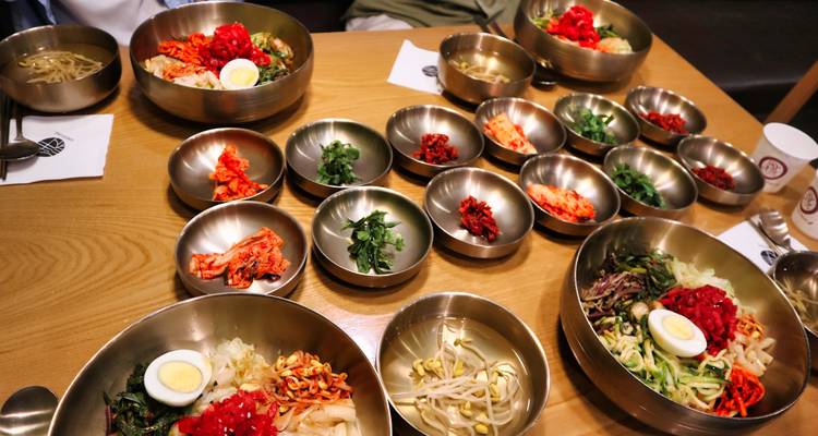 Una disposición de mesa de cuencos de latón llenos de ingredientes de bibimbap y acompañamientos coreanos espera a los comensales.