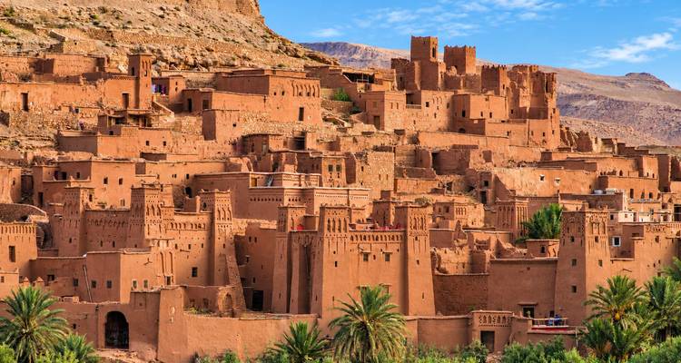 Los kasbahs de adobe de Ait Benhaddou se alzan majestuosamente contra un cielo azul brillante, con palmeras salpicando el primer plano.
