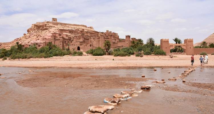 Una vista panorámica de Ait Benhaddou desde un lecho de río poco profundo con viajeros cruzando por las piedras de paso.