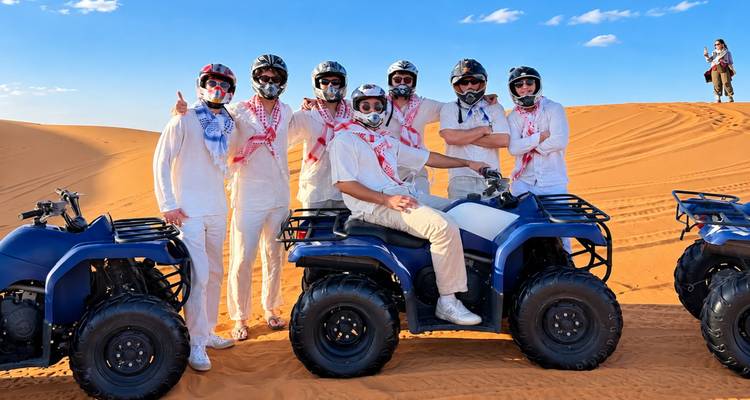Un grupo de amigos con trajes blancos posan en quads azules encima de las dunas doradas del Sahara bajo cielos despejados.