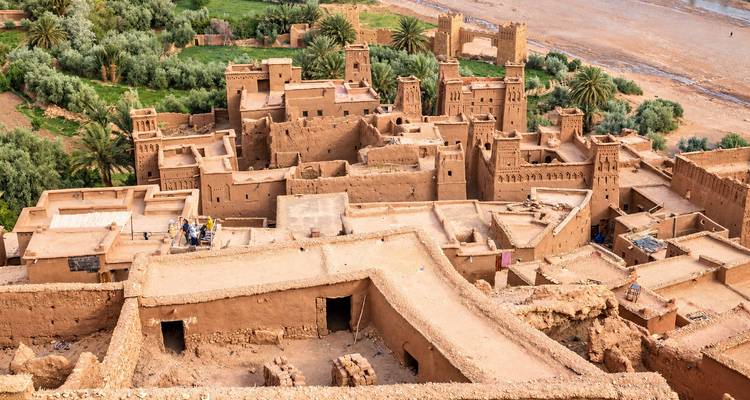 Een verhoogd uitzicht over de leemstenen torens en steegjes van Ait Benhaddou met weelderige rivierpalmen eronder.