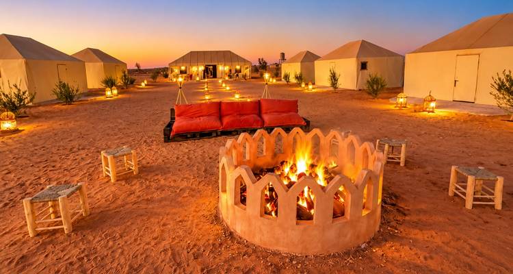Fogata central rodeada de tiendas iluminadas con linternas al atardecer en un campamento del desierto.