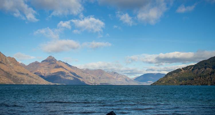 Lac bleu calme entouré de montagnes ondulées sous un ciel partiellement nuageux.