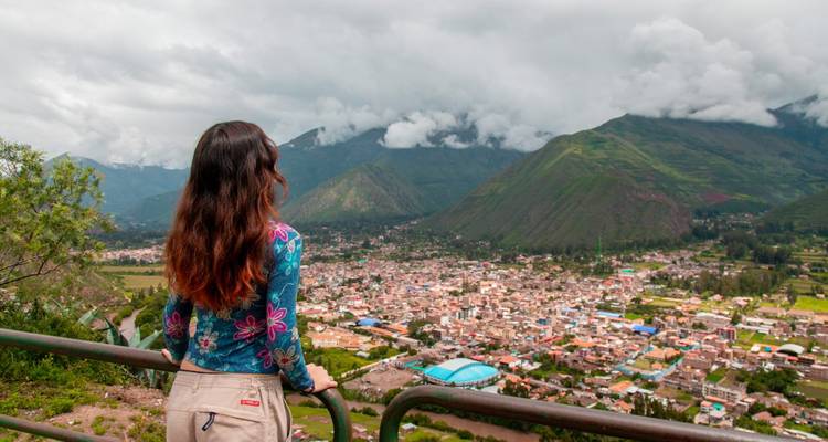 Une voyageuse contemple la ville étendue de la Vallée Sacrée et les montagnes luxuriantes sous un ciel nuageux spectaculaire.