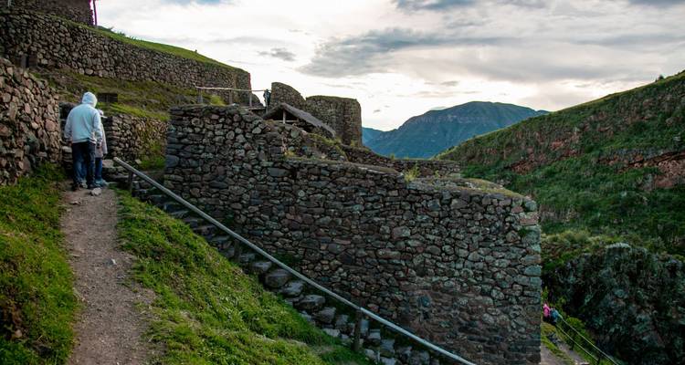Deux visiteurs marchent sur un étroit sentier de terre battue le long de ruines de pierre patinées qui surplombent les sommets andins au crépuscule.
