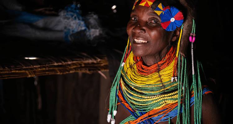 Femme âgée Himba à l'intérieur portant des colliers de perles colorés et des ornements de tête, éclairée en douceur sur un fond sombre