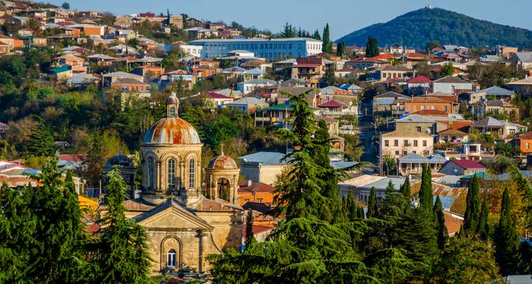 Paisaje urbano de una ciudad georgiana con una iglesia de piedra con cúpula en primer plano enmarcada por árboles perennes y casas coloridas en la ladera al fondo.