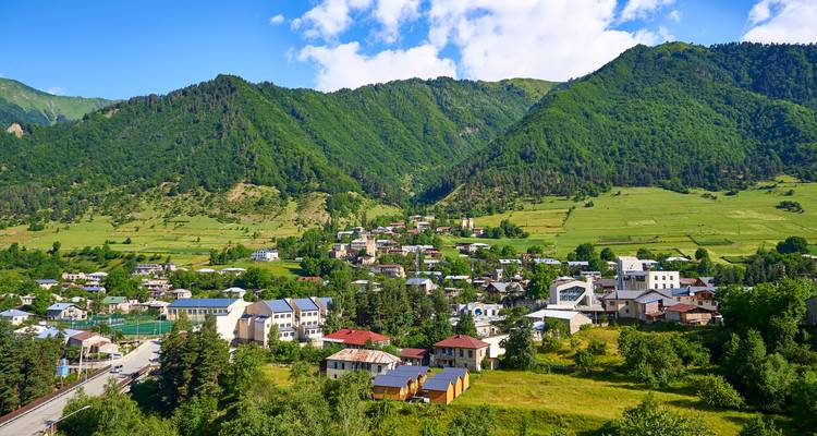 Asentamiento en valle montañoso con casas dispersas y laderas boscosas que se elevan hacia altos picos verdes bajo un cielo brillante.