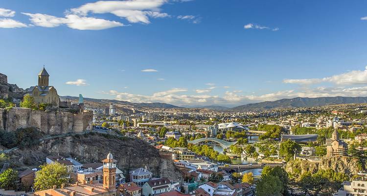 Vista panorámica de la ciudad de Tbilisi con la Fortaleza de Narikala y el moderno Puente de la Paz bajo cielos azules.