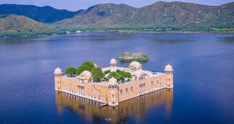 Vue aérienne du palais Jal Mahal flottant sur un lac bleu tranquille entouré de collines arides.