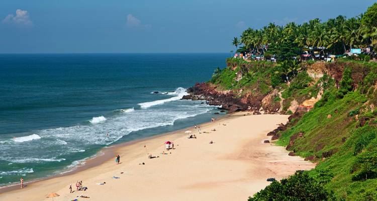 Goldener Sandstrand vor grünen Klippen und Palmen mit Sonnenanbetern entlang der Küste verstreut.