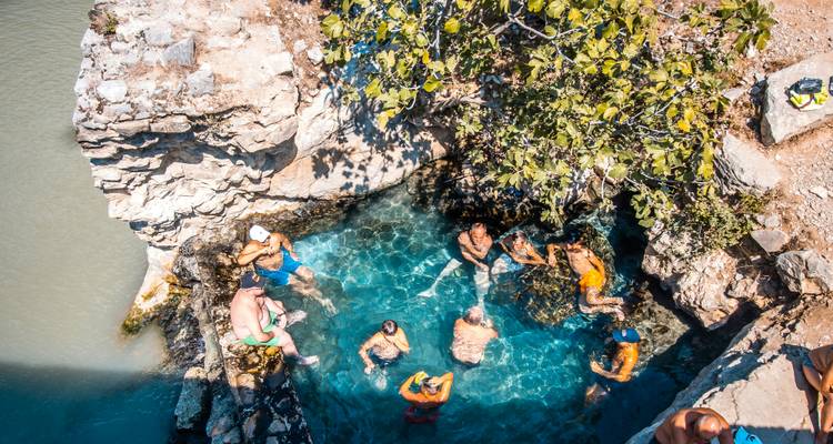 A dozen travelers soak in crystal-clear natural hot springs carved into rock beside a river