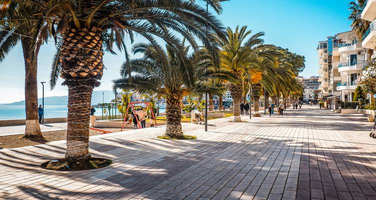 Palm-lined seaside promenade with locals strolling and playground equipment under bright sun