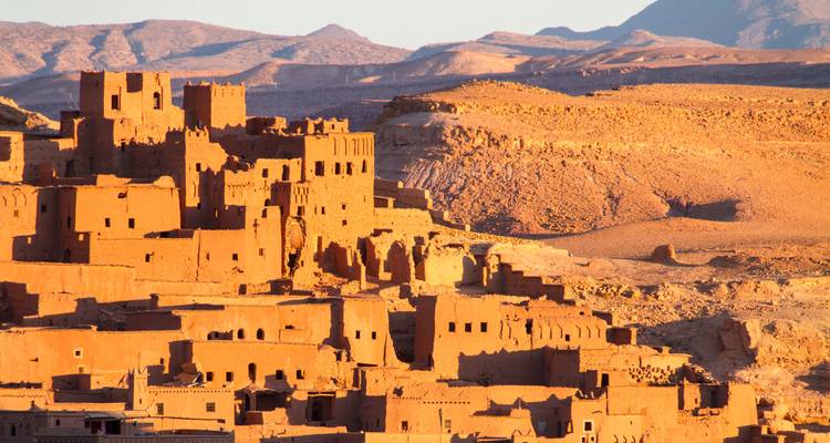 Les bâtiments en adobe aux teintes dorées d'Aït Benhaddou illuminés par le soleil de fin d'après-midi contre des collines escarpées.