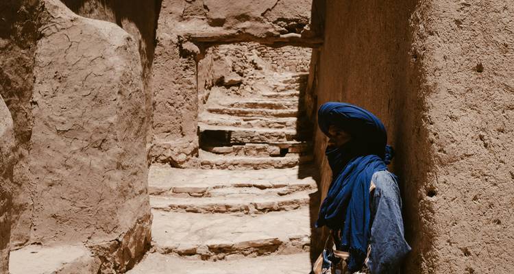 Un homme amazigh en robe indigo attend dans une ruelle en terre crue étroite et ensoleillée.