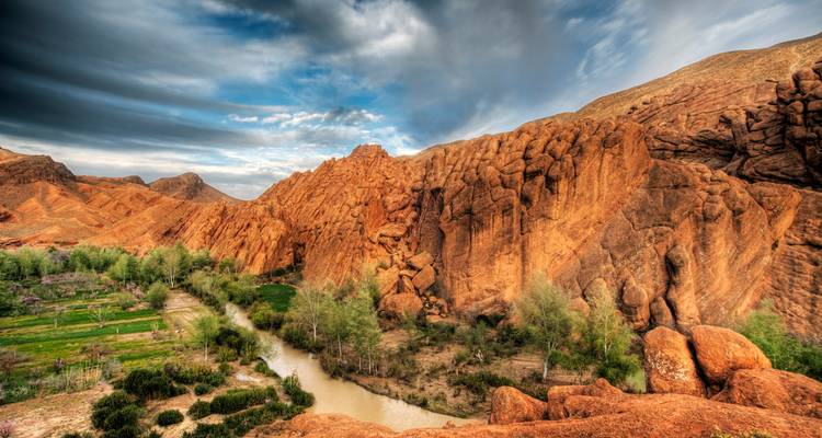 Les formations rocheuses rouges spectaculaires des Gorges du Dadès avec une rivière et des taches vertes sous un ciel chargé.