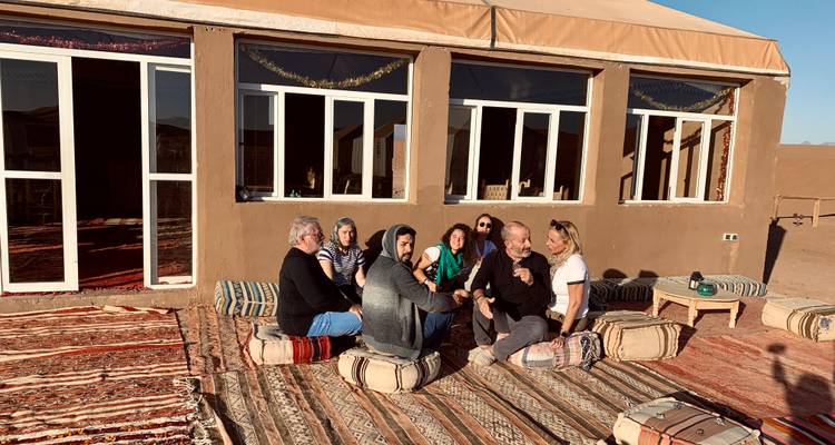 Des voyageurs assis sur des coussins dans un camp du désert à proximité d'un bâtiment de style adobe, discutant sous le soleil de l'après-midi.
