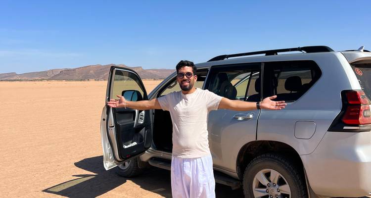 Voyageur souriant se tenant les bras écartés à côté d'un véhicule utilitaire sport sur une vaste plaine désertique aride.