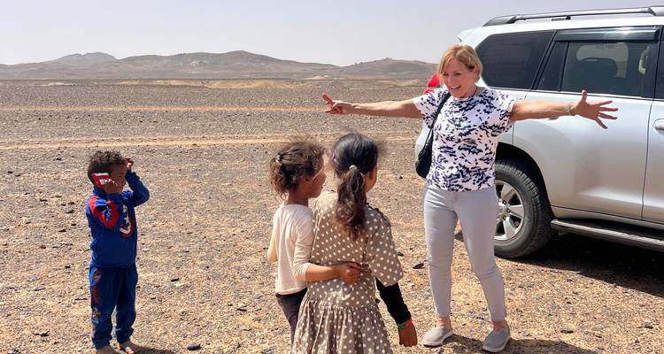 Une femme accueille joyeusement trois enfants du coin à côté d'un VUS dans une vaste plaine désertique rocheuse avec des collines basses en arrière-plan