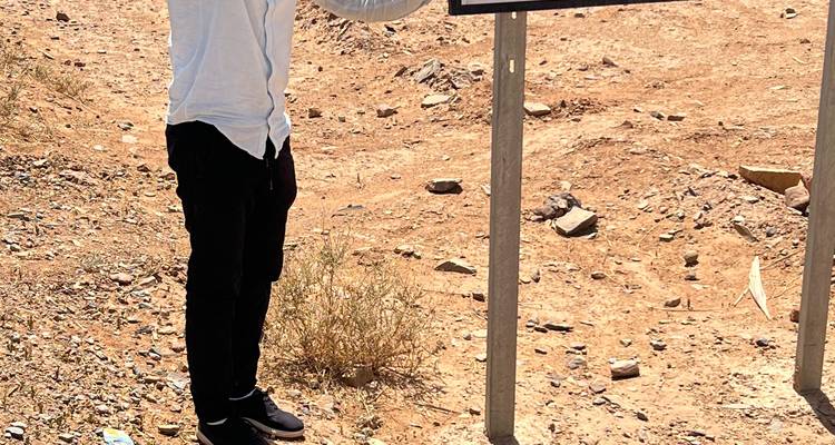 Un voyageur pose de manière ludique à côté d'une pancarte routière dans un paysage aride et poussiéreux.