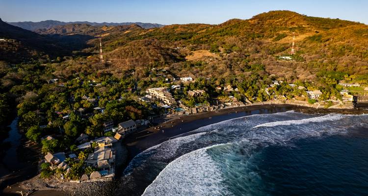 Vista aérea de un pequeño pueblo costero enclavado entre colinas boscosas y una playa curva de arena negra.