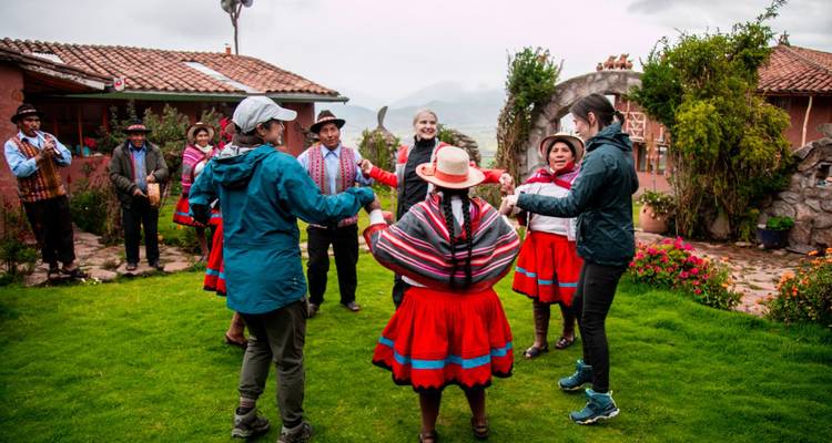 Des touristes et des femmes locales en tenue traditionnelle rouge se donnent la main en dansant sur une pelouse verte devant une maison de village