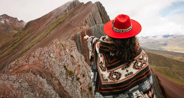 Excursionista con sombrero rojo y poncho estampado señala hacia la cresta rayada de la montaña de Perú.