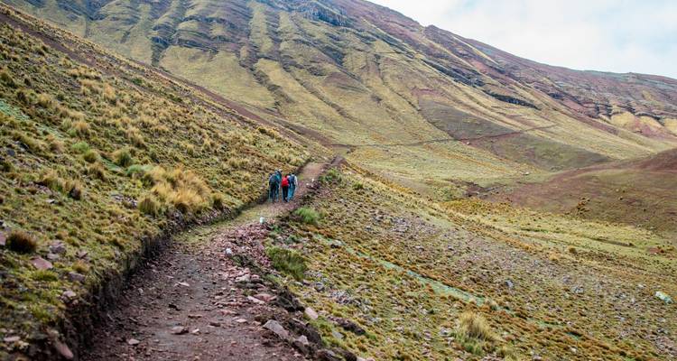 Dos excursionistas ascienden por un sendero rocoso a través de laderas andinas onduladas cubiertas de hierba.